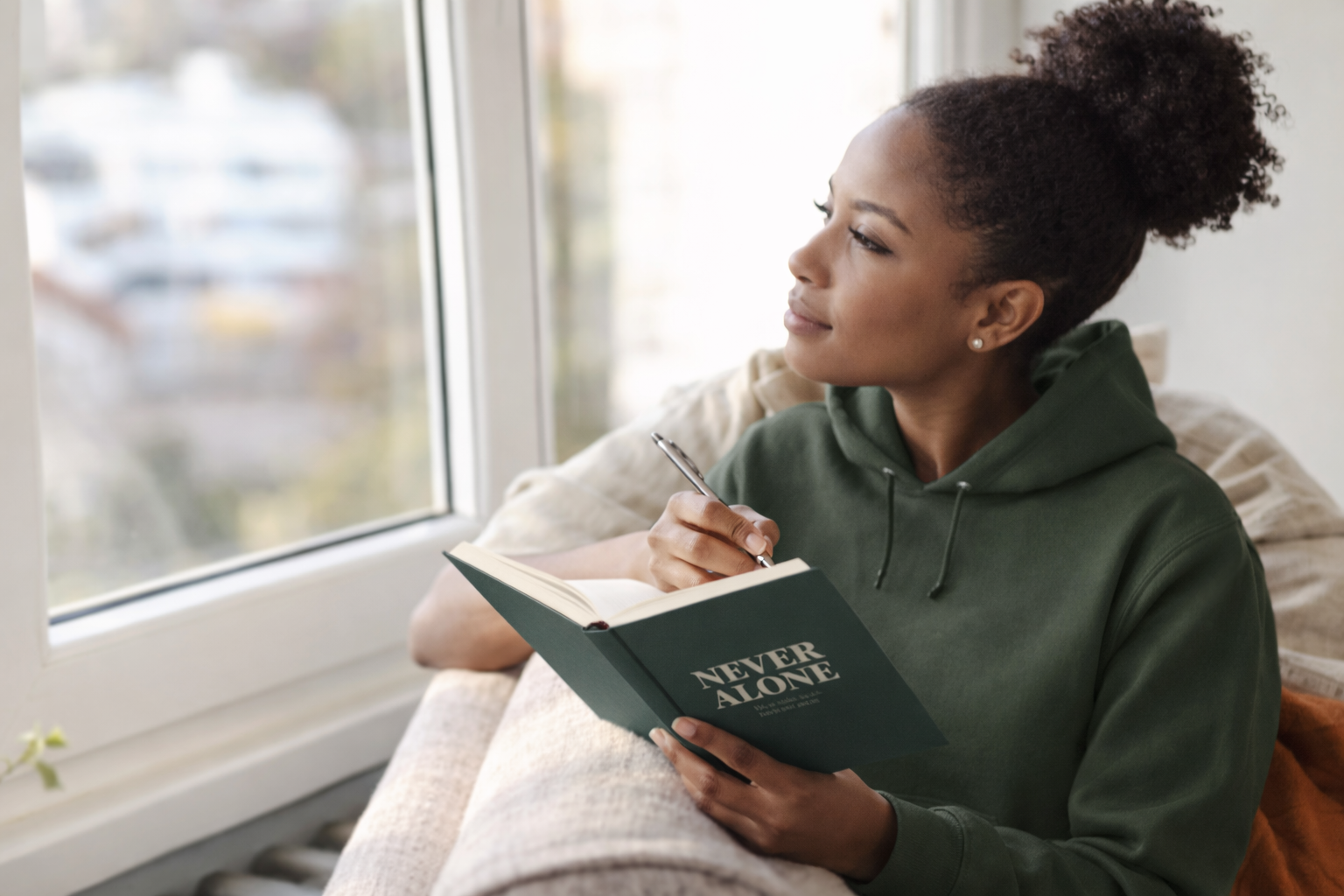 Woman in a green hoodie holding a journal called  'Never Alone' by a window.